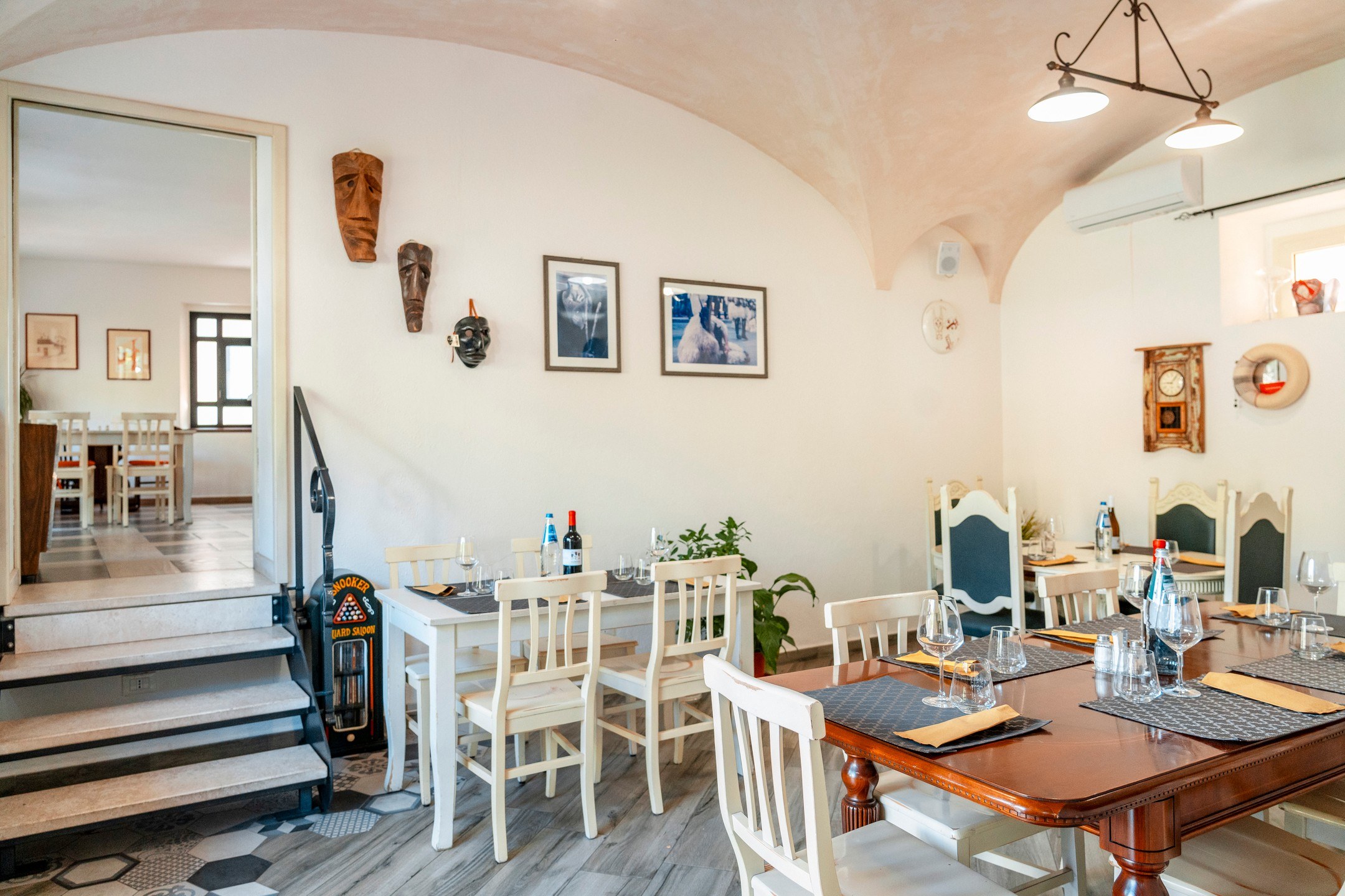 Sala di Manu Bar Trattoria con volta chiara, tavoli apparecchiati, sedie bianche e maschere in legno alle pareti
