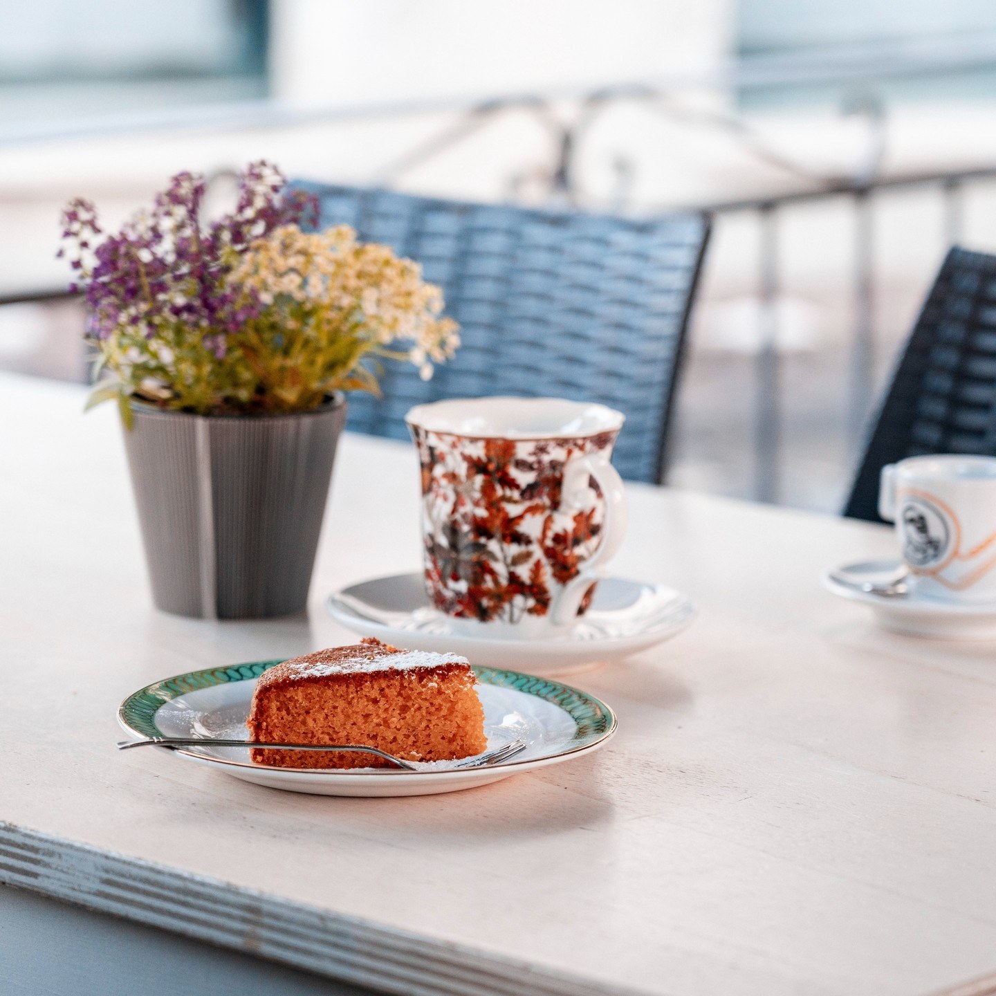 Fetta di torta con zucchero a velo su piattino bordato di verde, tazza a fiori rossi e vasetto di fiori su tavolo all'aperto