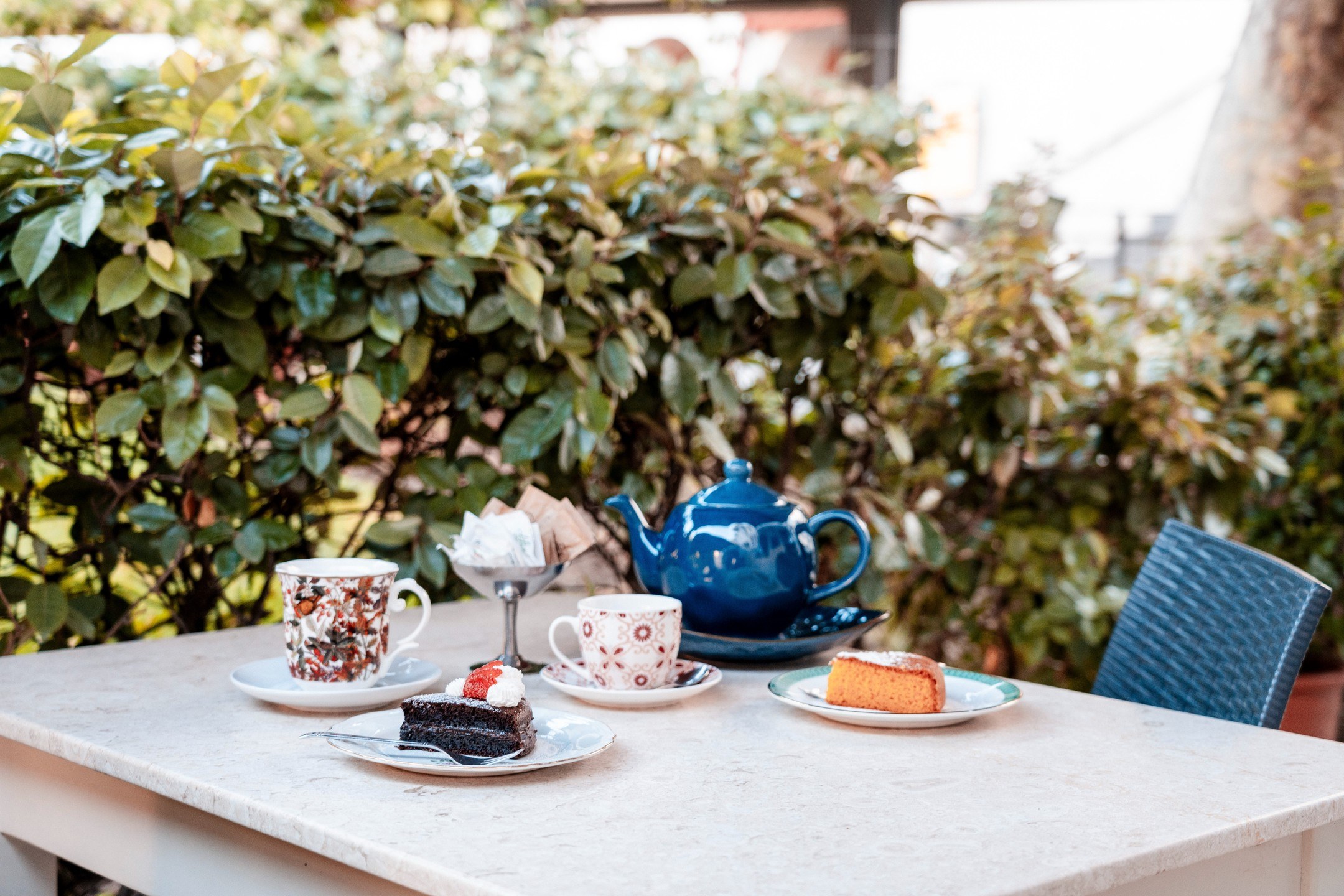 Teiera blu, tazze decorate e fette di torta al cioccolato e all'arancia su tavolo di marmo in giardino, Manu Bar Trattoria