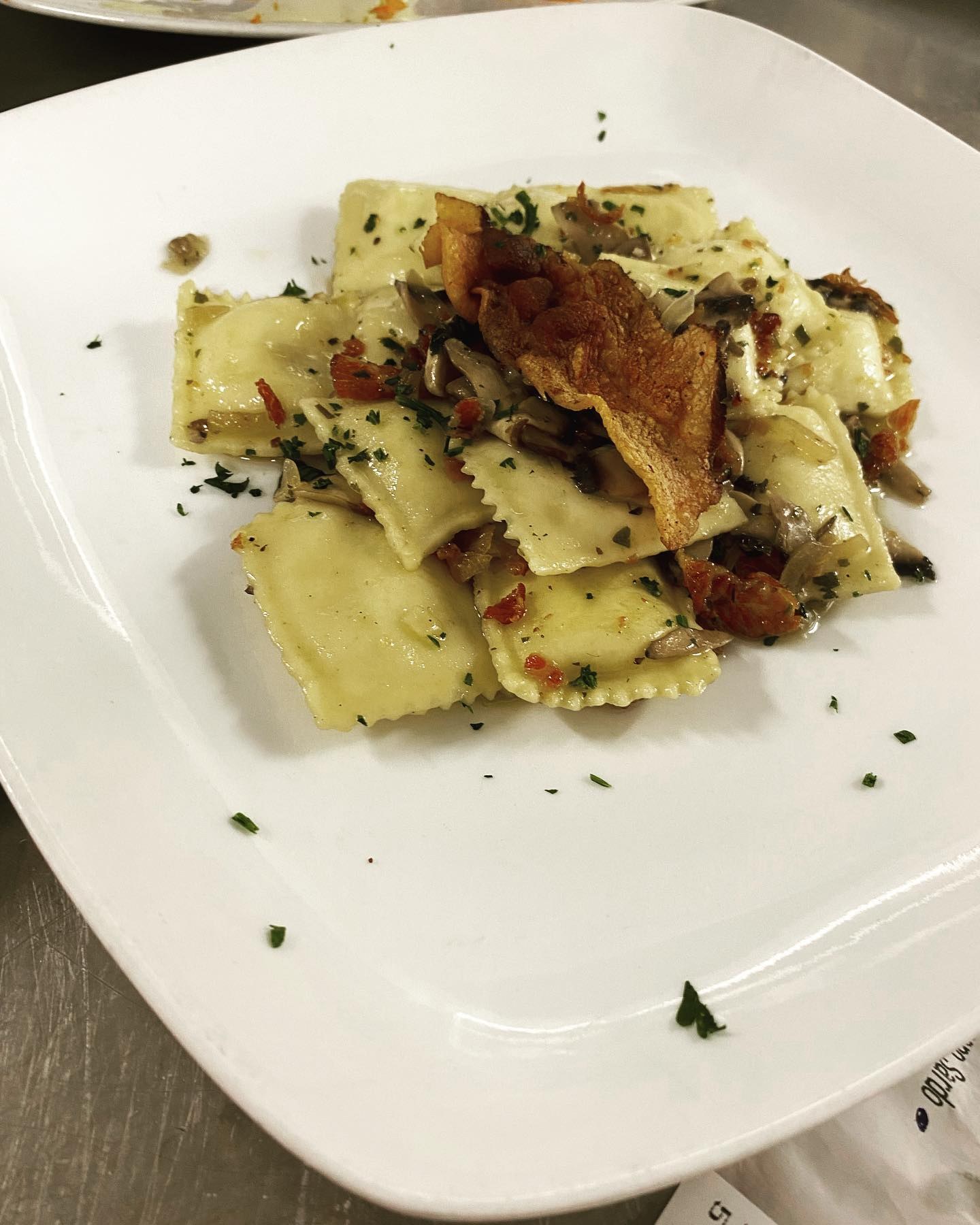 Ravioli con funghi, pancetta croccante e prezzemolo su piatto bianco quadrato, primo di Manu Bar Trattoria